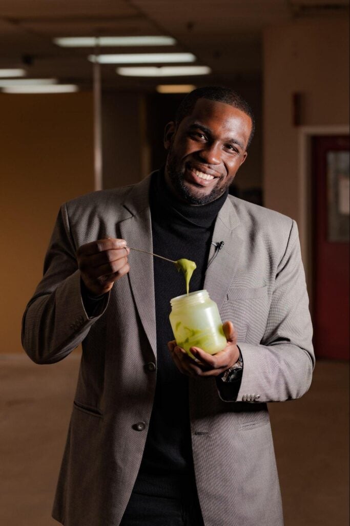 A man in a grey suit and black turtleneck smiles while holding a jar of green slime and lifting some with a stick. He stands indoors with blurred brown and red walls in the background.