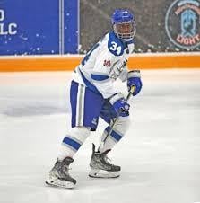 A hockey player in blue and white gear with the number 34 on their jersey skates on an ice rink, holding a hockey stick and looking ahead.