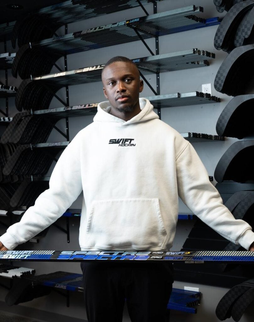 A man in a white hoodie stands indoors holding a hockey stick. Behind him, several shelves are lined with various hockey sticks, creating a sports equipment backdrop.