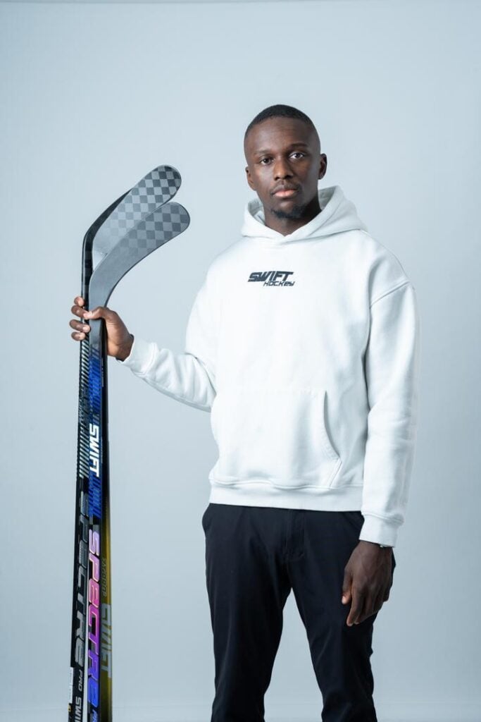 A man in a white hoodie and black pants stands holding two black hockey sticks upright against a plain light gray background.