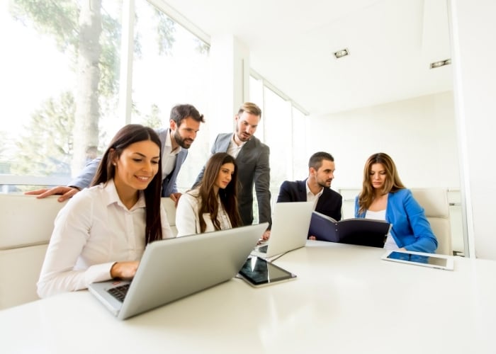Six business professionals are gathered around a white table in a bright office. Some are working on laptops, while others review a folder together. Large windows let in natural daylight.
