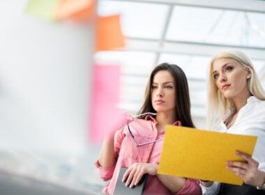 Two women stand indoors, focused on colorful sticky notes on a wall. One holds a folder and the other holds eyeglasses, both appearing to discuss or plan something in a bright, modern office space.