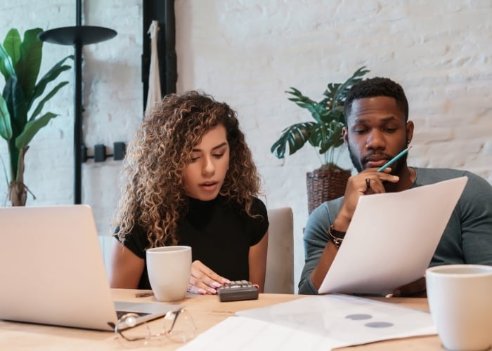Two people sit at a table with laptops, paperwork, and coffee mugs. The woman uses a calculator while the man looks thoughtfully at a document, appearing to discuss or review financial information.