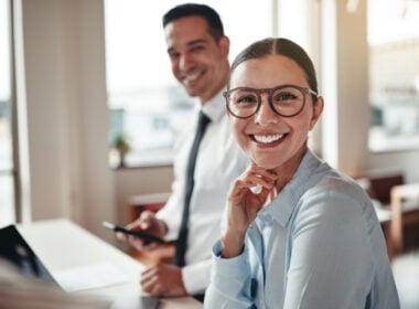 A woman wearing glasses and a light blue shirt smiles at the camera while sitting at a desk; a man in business attire stands blurred in the background, also smiling.