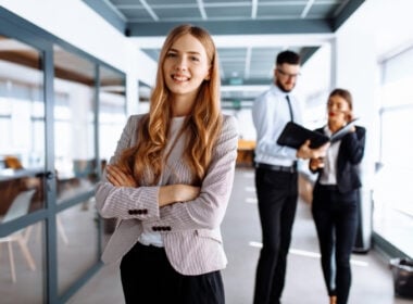 A confident young woman with long blonde hair stands with arms crossed in an office. Two colleagues are in the background, discussing paperwork. The setting is modern and professional.