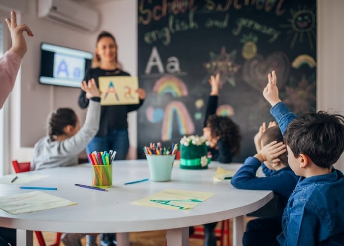 A teacher holds up a drawing of the letter A in front of a chalkboard as young children sitting at a table raise their hands in a classroom decorated with colorful art supplies and drawings.