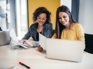 Two women sit at a desk, smiling as they look at papers and a laptop. One wears glasses and a denim jacket; the other wears a yellow top. Charts and pens are on the table in a bright, modern office setting.