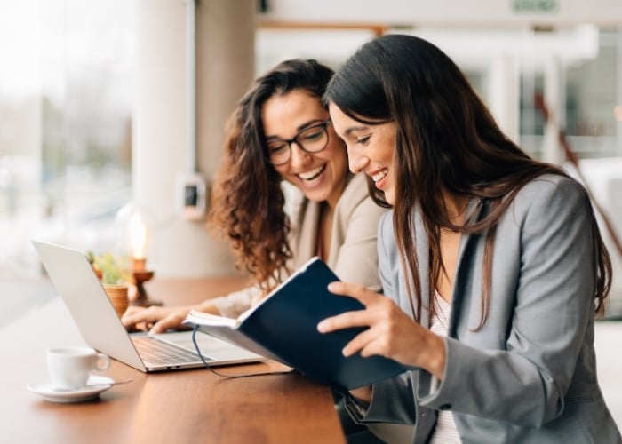 Two women in business attire sit at a table, smiling and working together on a laptop and notebook in a bright, modern office or café setting. A cup of coffee is on the table beside them.