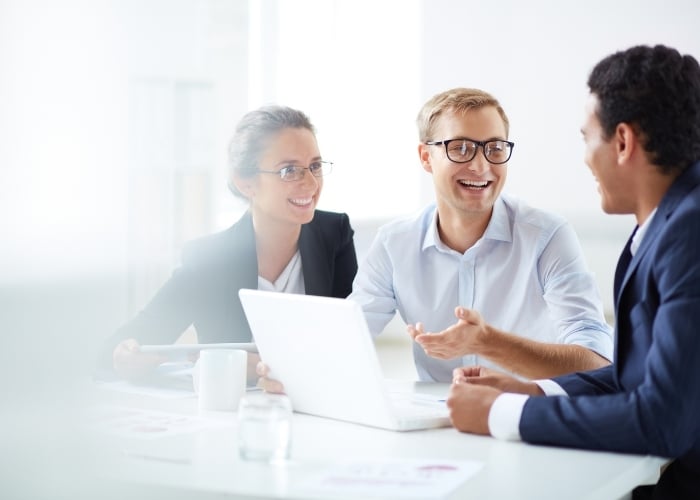 Three people in business attire sit at a table with a laptop, smiling and having a discussion in a bright office setting.