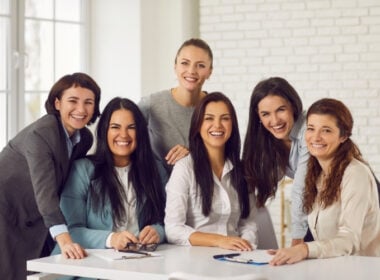 Six women dressed in business attire are smiling and gathered around a table in a bright office setting, suggesting a friendly and collaborative work environment.