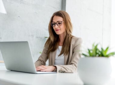 A woman wearing glasses and a beige blazer works on a laptop at a white desk in a modern office. A small green plant is in the foreground.