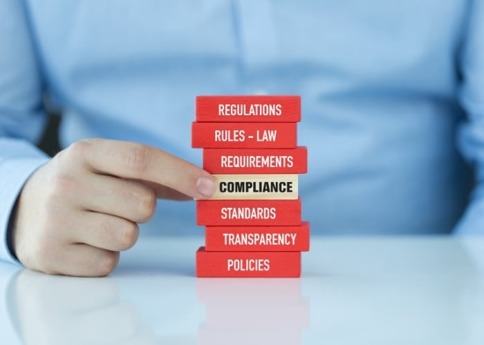 A person’s hand touches a stack of red blocks labeled with words like REGULATIONS, RULES–LAW, REQUIREMENTS, STANDARDS, TRANSPARENCY, and POLICIES. A central beige block is labeled COMPLIANCE.