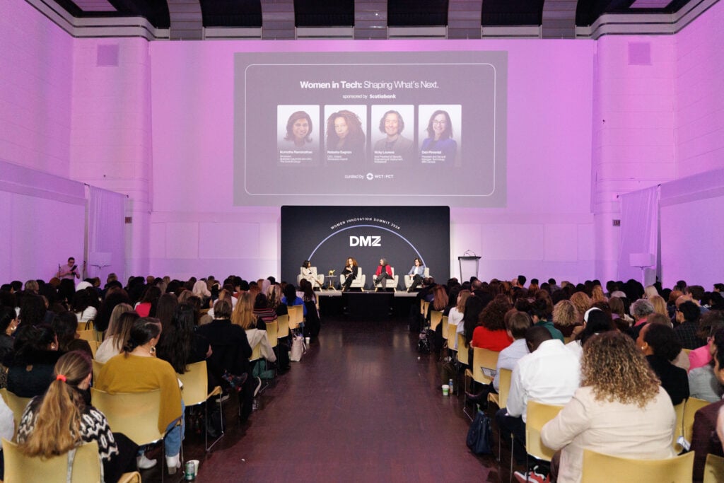 A large audience listens to a panel of five women discussing Women in Tech: Shaping What’s Next on a stage in a spacious, modern event hall with a DMZ sign and speaker photos projected on a screen behind them.
