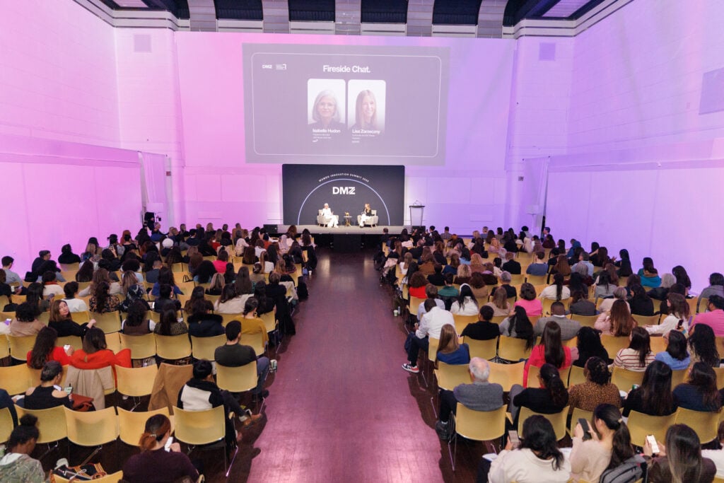 A large audience sits facing a stage where two people are seated for a fireside chat. A screen behind them displays their names and photos. The event space is brightly lit with purple accents.
