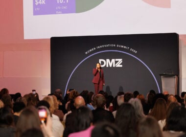 A speaker in a maroon suit addresses a seated audience at the DMZ Women Innovation Summit 2023, standing on stage in front of a black backdrop and a projected presentation slide.