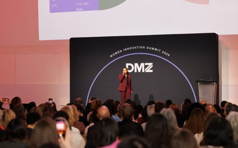 A speaker in a maroon suit addresses a seated audience at the DMZ Women Innovation Summit 2023, standing on stage in front of a black backdrop and a projected presentation slide.