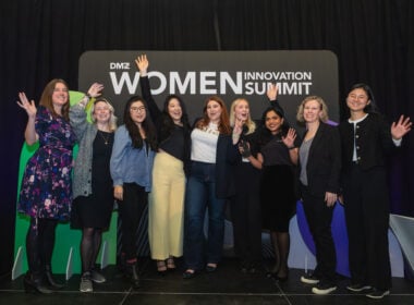 Ten women stand together on stage, smiling and waving, in front of a sign that reads “DMZ Women Innovation Summit.” They appear happy and celebratory, with a black curtain backdrop behind them.