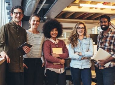 Five young adults, three women and two men, stand indoors in a modern office, smiling at the camera. Some hold notebooks or tablets. The group appears friendly and professional, with a casual, collaborative atmosphere.