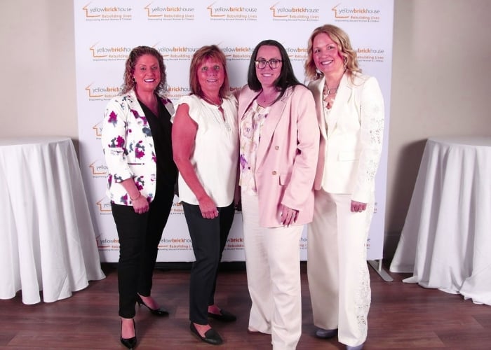 Four women pose together, dressed in business attire, in front of a step-and-repeat banner that reads yellowbrickhouse. Two round cocktail tables with white tablecloths are on either side of the group.