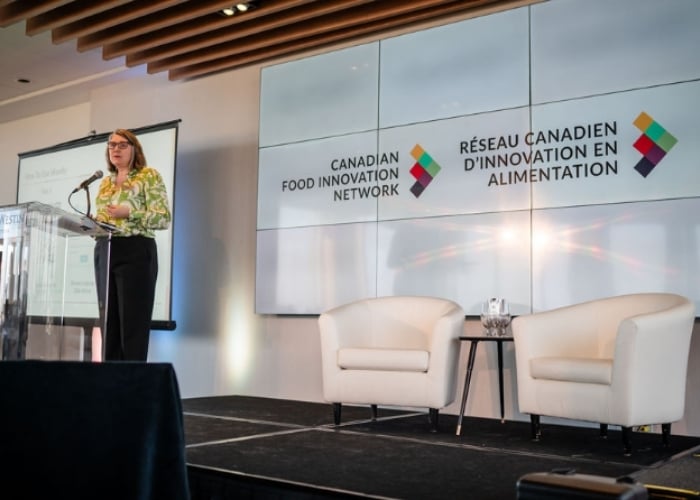 A woman stands at a podium giving a presentation on a stage with two empty white chairs. Behind her, large screens display the Canadian Food Innovation Network logo in English and French.