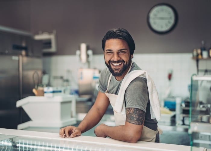 A smiling man wearing an apron stands behind the counter of a modern shop or café, leaning forward with one arm resting on the counter. A clock and kitchen equipment are visible in the background.