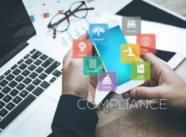 A person holds a smartphone near a laptop with charts, while colorful compliance-related icons and the word COMPLIANCE are overlaid, representing business standards, rules, and regulations.