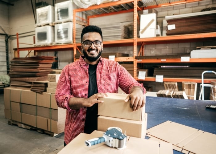 A smiling man wearing glasses and a red checkered shirt stands in a warehouse, holding cardboard boxes on a table with packing tape and stacked shelves in the background.