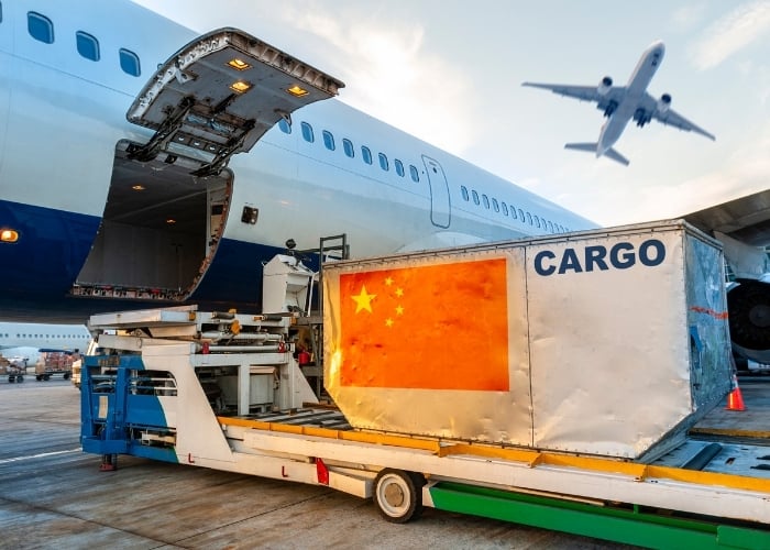 A large cargo container with the Chinese flag and the word CARGO is being loaded into an airplane, with another plane flying overhead at sunset on an airport tarmac.
