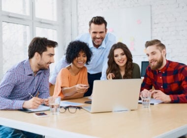 Five people sit and stand around a table, smiling and looking at a laptop screen. They appear to be collaborating in a bright, modern office setting with papers and glasses of water on the table.