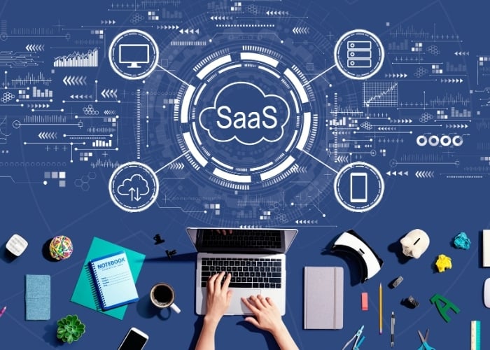 A person types on a laptop surrounded by office supplies, electronics, and SaaS (Software as a Service) icons connected by digital lines on a blue background with tech graphics.