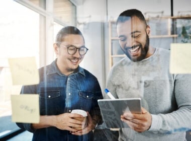Two men in casual clothes smile and look at a tablet together in an office, surrounded by sticky notes on a glass wall. One holds a coffee cup while the other holds the tablet and a marker.