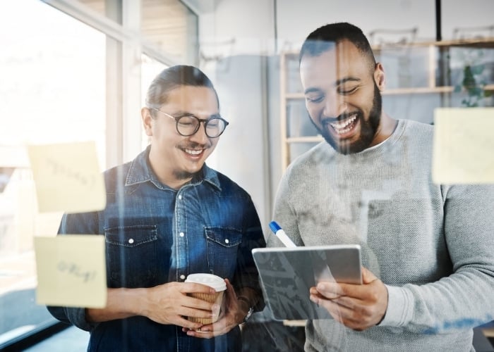 Two men in casual clothes smile and look at a tablet together in an office, surrounded by sticky notes on a glass wall. One holds a coffee cup while the other holds the tablet and a marker.