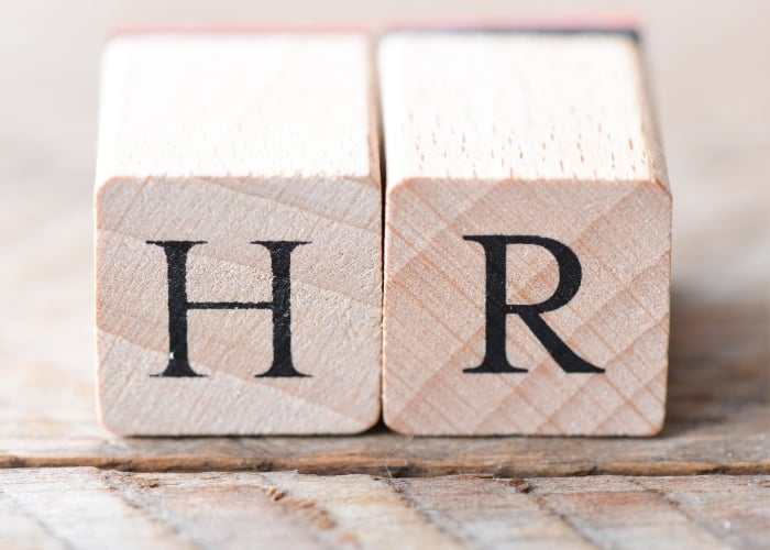 Two wooden blocks placed side by side display the black letters H and R, representing HR for Human Resources, on a wooden surface background.