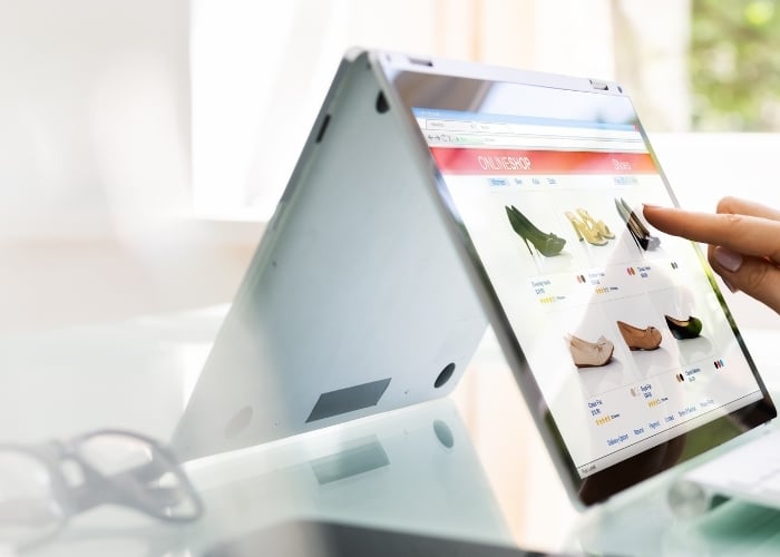 A person is using a touchscreen laptop in tent mode to browse an online shoe store, with images of various shoes displayed on the screen. The scene is bright and set near a window.