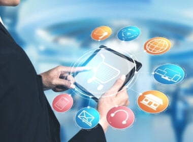 A person uses a tablet with a shopping cart icon on the screen, surrounded by colorful digital icons representing online shopping, communication, payment, and internet concepts, with a blurred blue background.