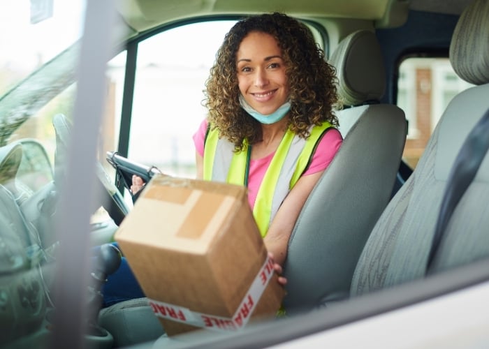 A woman wearing a reflective vest and a face mask under her chin sits in a vehicle’s driver seat, smiling and holding a cardboard box labeled FRAGILE.