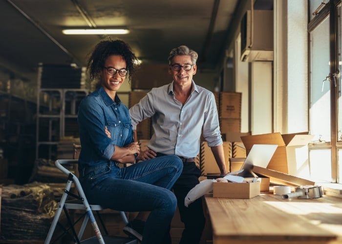 Two people, a woman and a man, smile while working together in a warehouse or office space with packing boxes, tape, and a laptop on a table beside them. Sunlight streams in through the window.