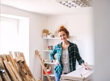 A smiling woman with curly red hair in a bun stands in a bright workshop, wearing a green plaid shirt over a gray t-shirt. She is surrounded by wooden planks and shelves with tools and supplies.