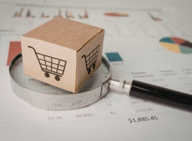 A cardboard box with a shopping cart symbol sits on a magnifying glass, placed over financial charts and graphs, suggesting analysis of online shopping or e-commerce data.
