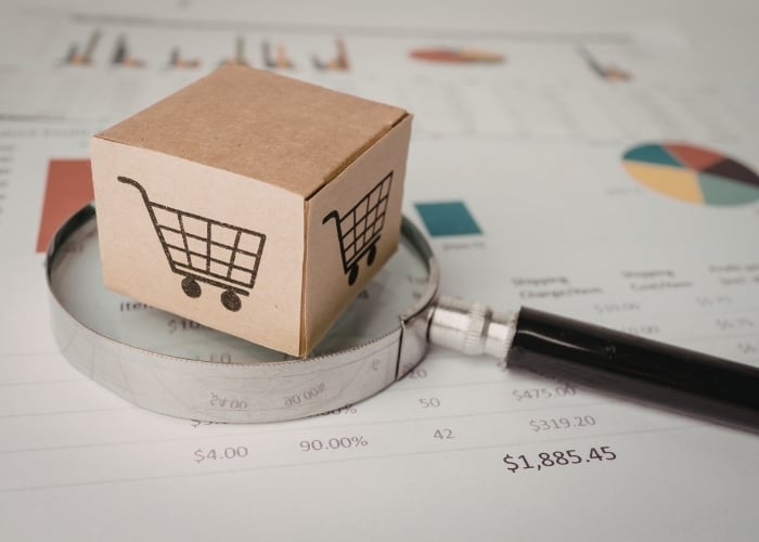 A cardboard box with a shopping cart symbol sits on a magnifying glass, placed over financial charts and graphs, suggesting analysis of online shopping or e-commerce data.