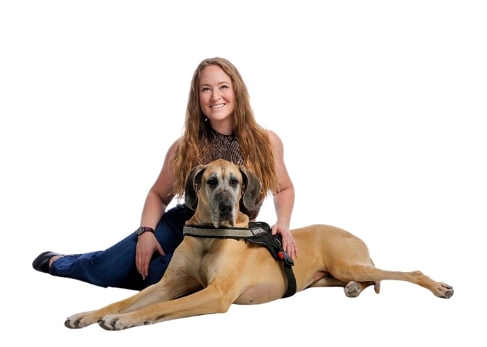A smiling woman with long hair sits on the floor behind a large tan Great Dane wearing a harness, both posing against a plain white background.