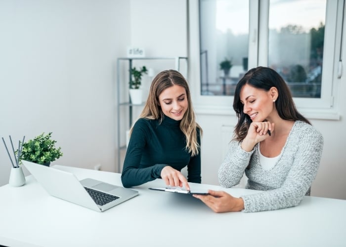 Two women sit at a white desk with a laptop and potted plants, smiling and looking at a clipboard together in a bright, modern office.