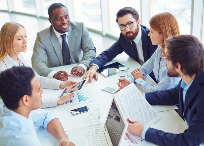 Six business professionals sit around a table, engaged in a discussion. Laptops, documents, and charts are spread across the table, and two people are holding a tablet and papers. The scene appears collaborative.