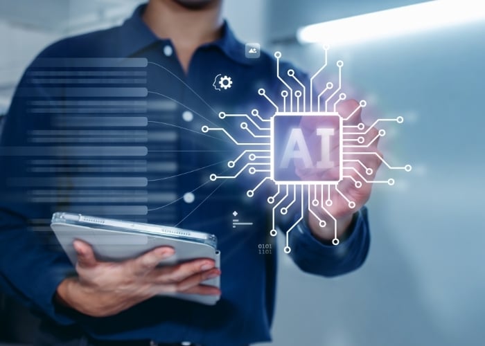 A person holding a tablet interacts with a glowing digital interface displaying the letters AI at the center, surrounded by circuitry graphics symbolizing artificial intelligence technology.