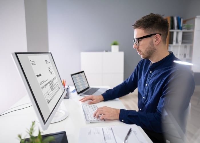 A man wearing glasses and a blue shirt works at a desk with a computer displaying an invoice. A laptop, papers, and pens are on the desk in a modern, bright office setting.