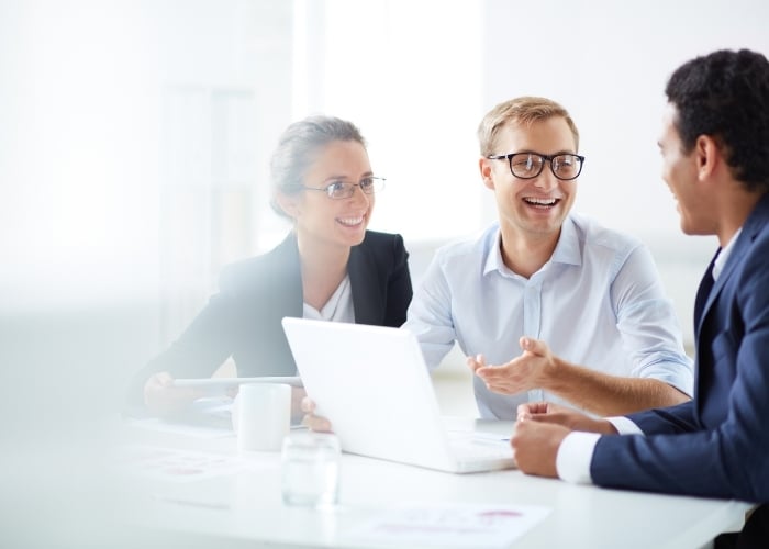 Three people in business attire sit at a table, smiling and talking, with a laptop and papers in front of them in a bright, modern office setting.