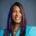 A woman with long, straight dark hair and purple highlights smiles at the camera. She is wearing a turquoise blazer, gold necklaces, and has a blue-grey background behind her.