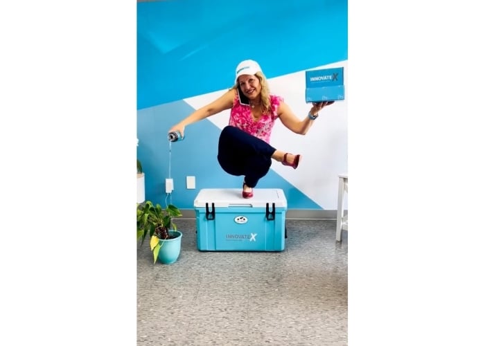 A smiling woman in a pink top and blue pants crouches on a blue cooler, pouring a drink from a can while holding a box. She wears a cap, and the background features a blue and white wall. A plant is beside her.
