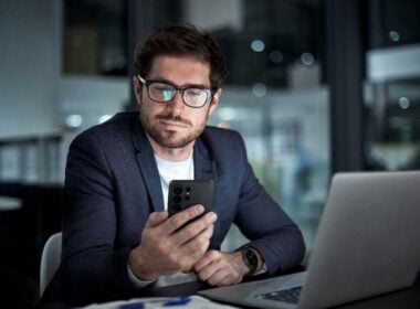 A man wearing glasses and a blazer sits at a desk with a laptop, looking at his smartphone in an office setting at night.