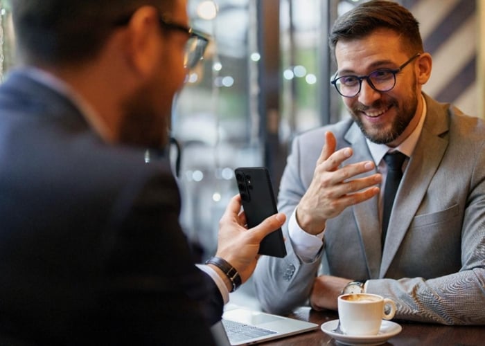Two men in suits sit at a café table talking. One holds a smartphone toward the other, who is smiling and gesturing with his hand. A cup of coffee and a laptop are on the table.